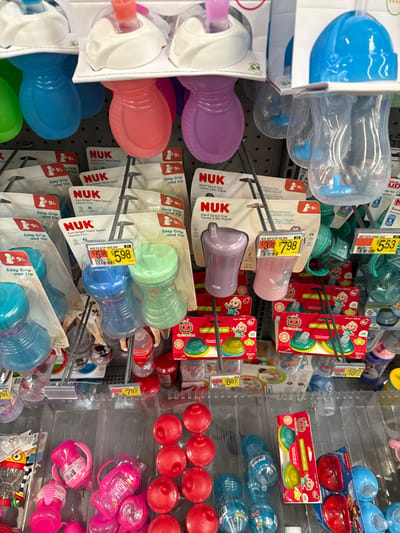 This image captures a brightly lit aisle in a retail store in Worcester, United States, showcasing a wide variety of baby bottles and sippy cups. The products are neatly displayed on shelves and hanging racks, indicating they are available for purchase. No people are visible in the scene.

On the left side of the image, numerous colorful NUK brand sippy cups are prominent, including "Hard Spout Cups" and "Easy Grip and Sip" models, in shades of blue, purple, pink, and green. Many of these are still in their transparent plastic packaging, featuring price tags such as "$5.98" and "$7.98." The packaging often displays a "9+" age recommendation.

Towards the center, additional baby feeding products, including smaller cups with cartoon characters, possibly from the "CoComelon" brand, are also hanging on display. Price tags like "$10.77" and "$8.87" are visible in this section. On the right, clear baby bottles with blue tops are stacked, alongside numerous red sippy cup bases arranged in rows. Below these, several pink sippy cups with handles are visible. A "CoComelon 3-Pack TIPPee" set is also displayed, with a price tag of "$7.97." Another price tag of "$5.53" is seen near some blue bottles at the top right.

The products are diverse in color and design, catering to different preferences and stages of infant development. The setting suggests a well-stocked baby care section within a supermarket or a large general merchandise store.