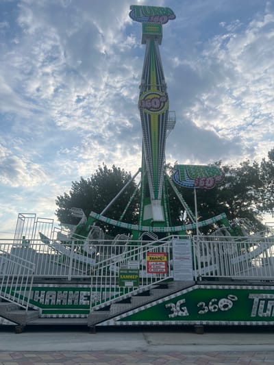 The image shows a large amusement park ride called "3G 360 Hammer" in Golubitskaya, Russia. The ride is green and white and features a tall, rotating arm with seating at the end. The ride is not currently in operation, and there are no people visible in the image. The sky is cloudy, and there are trees in the background. There are signs in Russian with information about the ride, including its name, height and time restrictions, and price. The ride appears to be located outdoors in an amusement park or fairground.