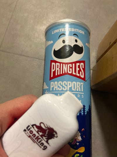 A photograph depicts a can of Pringles potato chips with a "Passport Flavors" label, lying horizontally on a grey tiled surface, next to a white bottle or container labeled with the Lion King logo being held in a human hand. The location is unknown; there is nothing in the image to indicate it is in Nanterre, France. There are also parts of a light brown wooden object visible.