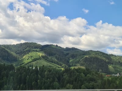 The image depicts a scenic view of lush, rolling green hills covered in dense conifer forests. The terrain shows varied slopes, with some areas appearing as clearings or meadows interspersed among the trees. The sky above is a vibrant blue, dotted with fluffy white clouds, suggesting fair weather. The lighting indicates it is daytime, with sunlight casting shadows and highlighting the contours of the landscape. In the distance, towards the lower right of the frame, there are hints of human settlement, including buildings with red roofs and what appear to be power lines or towers. The foreground shows the tops of trees, possibly from a closer vantage point, and a blurred white railing or barrier, suggesting the photo was taken from a moving vehicle or elevated position. The scene is characteristic of a mountainous or hilly region with significant forestation, and given the context, it represents a landscape in or around Judenburg, Austria.