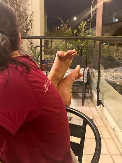 The scene depicts an individual seated on a residential balcony at night in Modiin-Maccabim-Reut, Israel. The subject, viewed from behind, wears a dark red short-sleeved shirt featuring light-colored Hebrew script, partially visible as "שלום." A gold-colored chain is present around their neck. The subject's bare feet are elevated and crossed, with soles visible, resting above a black metal chair armrest. The balcony features a glass and metal railing, light-colored tiled flooring, and several potted plants. In the background, outdoor lighting fixtures illuminate sparse vegetation against the dark night sky. Other furniture, including a chair and a patterned rug, are visible on the balcony floor.