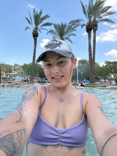 A woman in a purple swimsuit and baseball cap is taking a selfie in a body of water. She has tattoos on her chest and arm. Palm trees are visible behind her. In the background, people are gathered on the bank of the water. The location is in Emek HaMaayanot Regional Council, Israel.