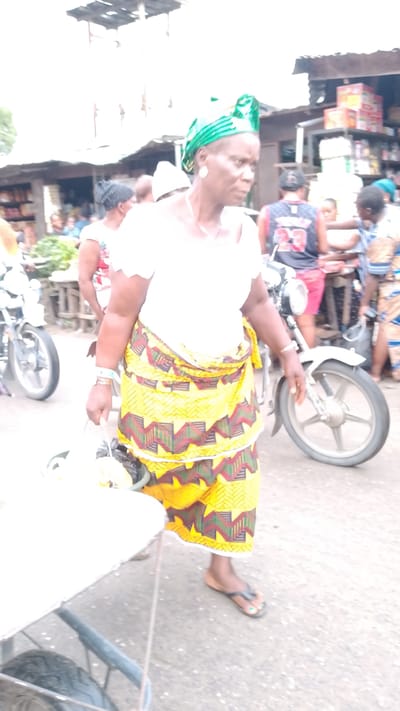 In Agbarho, Nigeria, a woman stands in an outdoor market setting, her gaze directed forward and slightly to the right. She wears a vibrant green headwrap adorned with metallic patterns and a white blouse with a lacy neckline. Her attire is completed by a yellow skirt featuring a geometric pattern of red, green, and brown zigzags and star shapes. She carries a light-colored bucket in her left hand, which appears to contain some items. Her right hand is by her side, with bracelets on her wrist. She is wearing black flip-flops. In the background, a white motorcycle with silver spokes is visible. Other individuals and market stalls are present, with goods displayed on shelves. The ground is paved, suggesting a market street or square. The lighting indicates it is daytime, with bright overhead light creating some overexposure.