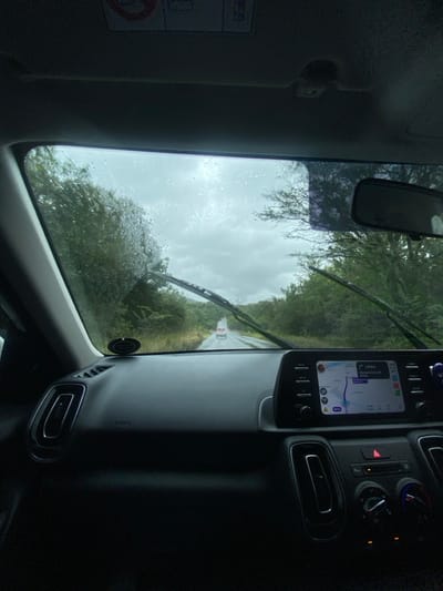 The image displays a first-person perspective from within a vehicle, looking out through the front windshield on a rainy, overcast day in Madibeng Nu, South Africa. The windshield is covered with numerous water droplets, partially obscuring the view. Two dark windshield wipers are visible in the lower section of the glass.

Outside the vehicle, a wet, asphalt road extends into the distance, flanked by dense green foliage and trees on both sides. A single light-colored vehicle is visible in the far distance on the road, traveling away from the viewer. The sky is uniformly grey and overcast.

Inside the vehicle, the dark dashboard occupies the lower portion of the frame. On the center console, a digital infotainment screen displays a navigation map, showing a route with an estimated 1.8 km distance remaining and the text "Madalelburg Nu." Below the screen, a red triangular hazard light button is illuminated, surrounded by other dark climate control buttons and knobs. Rectangular air vents are visible on the left side of the dashboard and below the infotainment screen. An "AIRBAG" label is printed on the passenger side dashboard. A rearview mirror is visible at the top center of the windshield, and a portion of a sun visor with an "AIRBAG" warning label is visible at the upper left. No individuals are present in the visible scene.