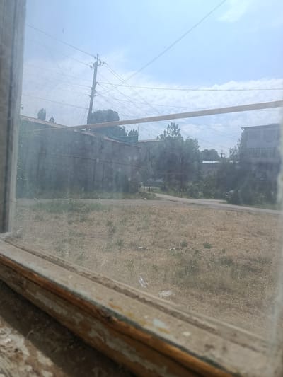 A daytime, outdoor, medium-wide shot captures a view through a dusty, weathered window frame of a rural scene. The foreground shows the aged wooden window sill and frame, with chipped paint and visible wear. Beyond the window, a dry, grassy field dominates the mid-ground. A dirt road runs across the field, leading towards buildings in the background.
In the distance, a row of industrial-looking buildings with corrugated metal roofs are visible, interspersed with trees. Further back, on the right side of the frame, a multi-story apartment building with a light-colored facade and numerous windows stands out. Another similar building is visible between the trees. Utility poles with a complex network of wires crisscross the sky, indicating the presence of electricity and telecommunications infrastructure. The sky is a pale blue, with scattered white clouds. The overall impression is one of a sunny, perhaps hot day in a somewhat neglected urban or semi-urban environment. There is no discernible activity or people in the frame, and no visible text. The scene appears to be located in Khujakent, Uzbekistan, as indicated by the location context.
