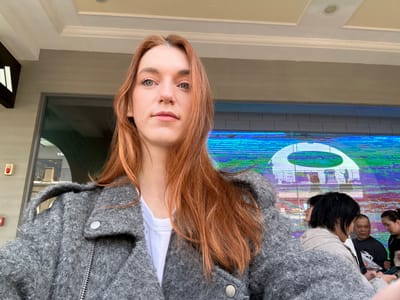 A fair-skinned woman with red hair and blue eyes looks directly at the camera. She wears a white shirt and a gray wool jacket. Behind her, a group of people are reflected in a glass wall with a large, circular logo. A storefront and various building details are visible, suggesting an urban setting. The image appears rotated.