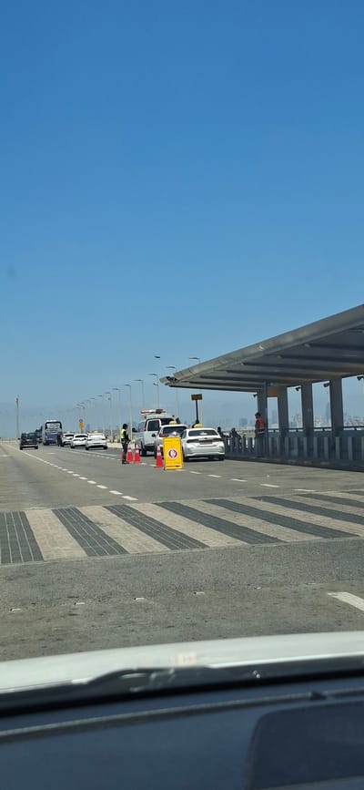 This image depicts a checkpoint on a road, possibly at an airport or a border crossing, in the city of Unknown, Israel. The sky is clear and blue. In the foreground, a car's dashboard is visible. The road has zebra crossings with black and white stripes.
Several vehicles are lined up: a black car, a blue bus, and multiple white cars, some of which appear to be taxis based on markings. There are also two white police or security vehicles.
Two individuals in uniform, likely security personnel, are present. One is standing near a police car, and another is to the left of the lane, wearing a yellow vest and holding a traffic cone. A yellow sign with red markings and text in Hebrew is positioned in the road, along with orange traffic cones, indicating a stop or restricted area.
To the right, a covered walkway or structure with a metal roof supported by columns runs parallel to the road. People are visible standing under this structure. In the background, a cityscape with buildings can be seen in the distance. Streetlights line the road. The overall scene suggests a controlled entry point.
