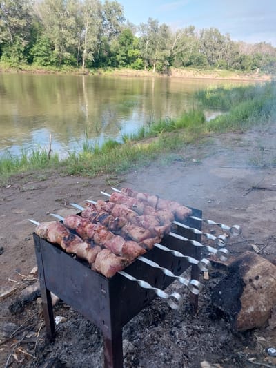 In an outdoor setting near the city of Chkalov, Russia, a metal barbecue grill stands on the ground, filled with skewers of raw meat. The grill is situated on a dirt and ash-covered surface, with remnants of a fire including burnt logs and wood. In the background, a wide, calm river reflects the sky and the lush green trees lining its banks. The sky is partly cloudy. The scene appears to be a picnic or a camping trip by the river.