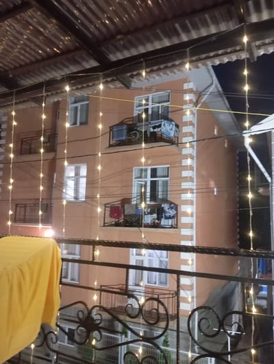 The image depicts the exterior of a multi-story building at night, adorned with strings of warm-toned fairy lights. The building has an orange facade with white decorative trim around the windows and corners. Several balconies are visible, some with railings and clothes hanging on them. The roof appears to be corrugated metal. The foreground shows a metal railing and a yellow cloth draped over an object. The scene is consistent with an urban residential setting in Bosteri, Kyrgyzstan, illuminated by decorative lighting after dark.