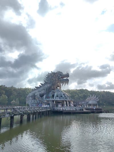 The image displays an abandoned dragon-themed structure situated on a body of water in Kim Long Ward, Vietnam. The central subject is a large, weathered grey dragon sculpture, exhibiting signs of deterioration and extensive graffiti, positioned atop and integrated with a circular, dome-roofed building. The dragon features an open mouth with visible teeth, distinct scales, and dorsal spikes. A concrete bridge, supported by pillars and showing wear, extends from the left foreground towards the structure, connecting it to land. Several individuals are observed walking or standing on this bridge and around the base of the main structure. The water in the foreground and midground is a calm, dark hue with ripples on its surface. A dense line of green trees borders the far side of the water, forming the background. The sky above is largely overcast with a mix of grey and white clouds.