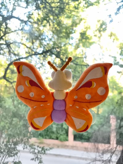 A plush toy butterfly with orange wings, a white body, and purple abdomen is attached to a window. The background is out of focus, showing green foliage and bright sunlight, suggesting an outdoor setting with trees. The specific location of Sokolivske, Ukraine, is not discernible from the image itself.