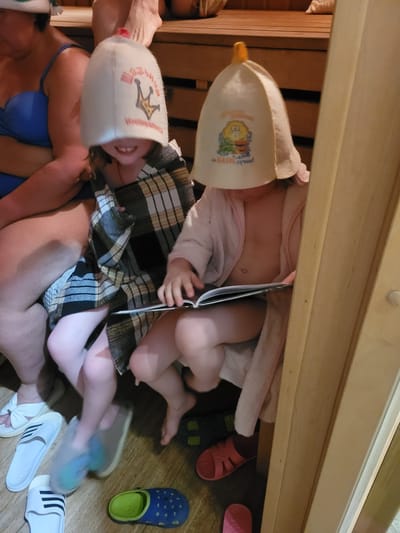 The image shows a group of people inside what appears to be a sauna. Two individuals, likely children, are positioned on a bench or platform, each wearing a sauna hat. One child is reading a book. A woman in a blue bikini and a man are also present, their legs and torsos visible at the bottom of the frame. Several pairs of sandals and shoes are scattered on the floor. The location is Пятрышкаўскі сельскі Савет, Belarus.