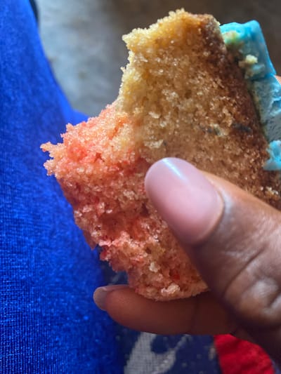 The image shows a close-up of a person's hand holding a piece of cake with blue frosting and pink cake. The person's hand is dark-skinned, and their fingernail is visible and appears to be manicured with a pinkish hue. The cake appears to be a slice from a larger cake, with a crumbly texture. The background is blurred, but it appears to be a blue fabric. The image does not provide any information about the location in the city of Wumat, Nigeria.