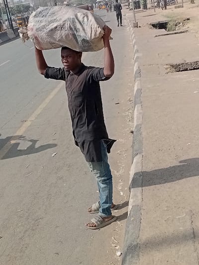 The image depicts a daytime scene on a street in Sharada, Nigeria. A young adult male, positioned left of center, is standing on the paved roadside next to a concrete curb. He is wearing a dark, long-sleeved tunic, faded blue jeans, and light-colored slide sandals. He balances a large, off-white, irregularly shaped sack on his head, supporting it with both arms raised. His gaze is directed forward.
The foreground and midground consist of a worn asphalt or concrete road surface with visible debris. A concrete curb separates the road from a dirt shoulder on the right, which contains an open drainage ditch or culvert section and additional scattered debris. In the background, a multi-lane road extends into the distance, where several individuals, "keke napep" (tuk-tuks), and other vehicles are present. Strong shadows cast by the curb and unseen objects indicate bright daylight conditions.