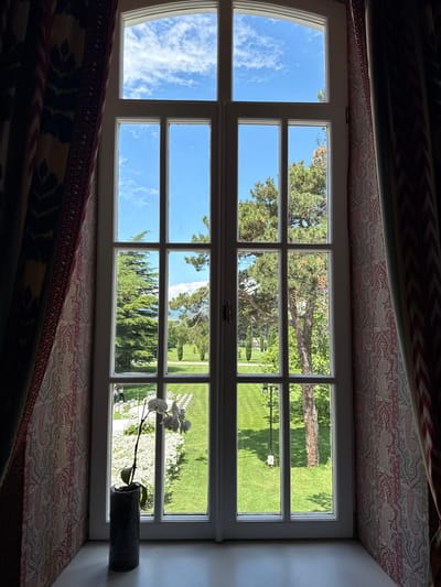 The image shows a view through a tall, multi-paned window looking out onto a garden. The window frame is white, and the panes are divided into a grid. On the left side of the window, a dark curtain with a patterned design hangs. On the right, another curtain with a lighter, repeating paisley pattern is visible.
In the foreground, on the windowsill, a tall, dark cylindrical vase holds a single orchid plant with white flowers and green leaves.
Through the window, a well-maintained garden is visible. The garden features a lush green lawn, mature trees, and white flowers in planters. In the middle ground, rows of white chairs are set up on the lawn, suggesting an event is planned or has recently occurred. Two people, appearing to be casually dressed, are standing near the chairs. Further in the distance, more trees and open grassy areas are visible, with a glimpse of distant hills or mountains under a bright blue sky with scattered white clouds. The overall impression is one of a peaceful, sunlit day in a well-kept outdoor space. This scene is located in Mukhrani, Georgia.