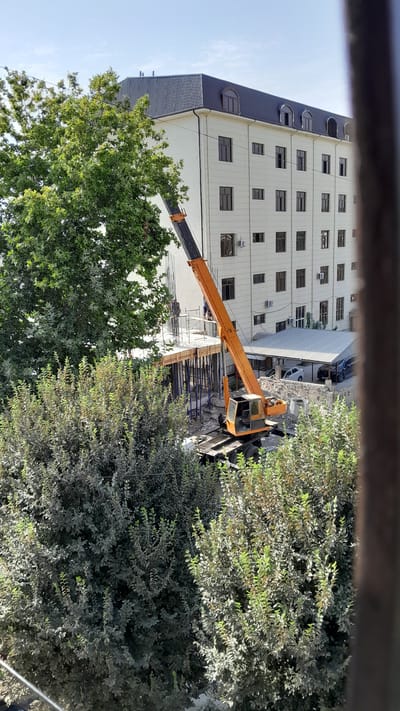 In Denov, Uzbekistan, a construction site is visible. An orange crane is positioned near a multi-story white building. The building has a dark, pitched roof with dormer windows. In the foreground and to the left of the building, large, mature trees with dense green foliage are present. The construction area itself features concrete formwork and structural supports. A few people are visible on the construction platform. The scene is set against a clear sky.