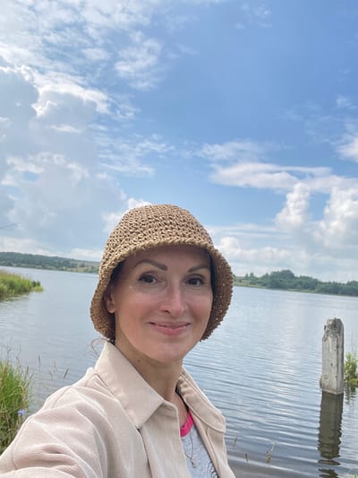 A woman is taking a selfie outdoors, in the city of Bereznik, Russia. She is wearing a straw hat and a light-colored corduroy jacket over a pink and grey shirt. She has brown hair and is smiling at the camera. She is standing by a lake, with trees and hills in the background under a cloudy blue sky. A concrete post is visible in the water to the right of the frame.