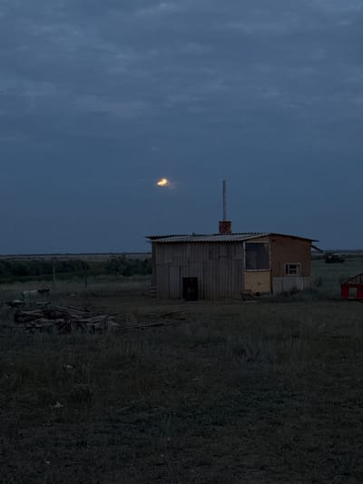 The image captures a dimly lit outdoor scene, likely in the evening or early morning given the muted sky and overcast conditions. The primary subject is a small, rustic wooden structure, possibly a shed or outbuilding, constructed with corrugated metal and wood panels. A chimney extends from its roof, and there are visible windows and a doorway. To the left of the structure, a small, bright object hangs in the sky, appearing as a streak of light or a distant, illuminated phenomenon.
The setting appears to be rural, with dry, tall grass covering the ground around the building. In the background, the terrain is flat and extends towards a distant, hazy horizon. There are also glimpses of other objects, including what seems to be a red agricultural vehicle or trailer further in the distance. The overall mood is somber and quiet, typical of twilight in a remote, agricultural region. No people are visible. There is no visible text in the image.
