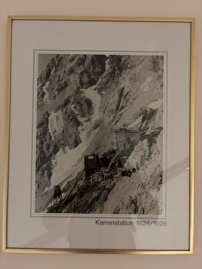 This is a framed black and white photograph depicting a mountain landscape with structures and people. The text at the bottom of the photograph reads "Kammstation 1924/1926," indicating it is a view of the Kammstation (summit station) from the years 1924-1926. The image shows a steep, rocky mountainside with patches of snow. Several wooden structures and what appear to be construction elements, possibly part of a cable car system or building, are visible on the slope. A group of people, small in number and scale, are gathered near some of the structures, suggesting human activity or construction work. The overall scene is rugged and mountainous, with evidence of human intervention in the form of the built structures. The photograph is matted with a light-colored border and framed with a thin gold-colored frame. The location is identified as the Kammstation in Ehrwald, Austria.
