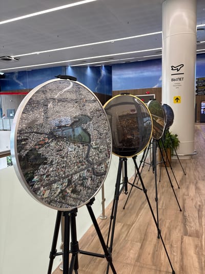 The image shows an art installation in what appears to be an airport or public transit hub, with a display of circular artworks mounted on tripods. The artworks are aerial photographs of urban landscapes. The backdrop is a mural of a sky with clouds, and a white pillar with signage indicating "DEPARTURES" in English, Russian, and Chinese. To the right of the pillar, there is another sign with vertical text that is not legible. The floor is made of light-colored wood planks. The overall environment suggests a modern, clean, and public space. The specific location of Старое Мартьяново, Russia is not indicated by any visual cues in the image.