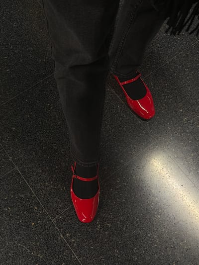 A downward shot features two legs in dark denim pants, ending with shiny red Mary Jane shoes over black socks. The floor is a dark, speckled tile, with a concentrated reflection of light near the upper right. A bit of black clothing is visible near the upper left. The focus highlights the contrast between the bright shoes and the muted surroundings.