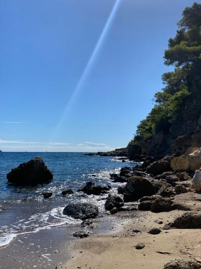 The image depicts a sunlit coastal scene in Megalochori, Greece. The foreground features a narrow, light brown sandy and pebbly beach strewn with numerous dark, irregular rocks of varying sizes. Small white-capped waves lap at the shore, creating white foam around the partially submerged rocks. To the left, a large, dark boulder rises from the water in the mid-ground. The deep blue sea extends across the middle of the frame, sparkling with sunlight reflections. Several faint white specks, likely distant sailboats, are visible on the horizon. The right side of the image is dominated by a steep, rocky cliff face, heavily vegetated with dense green trees, extending from the water's edge upwards to the frame's top right corner. The sky above is clear and brilliant blue, traversed by a strong, diagonal beam of sunlight or lens flare emanating from the upper left. No people or animals are present.
