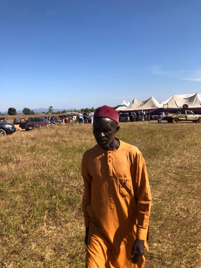 An outdoor image shows a man wearing a maroon cap and an orange button-up shirt. He stands on a grassy field on a sunny day with a clear blue sky. In the background, there are tents, a crowd of people, and parked vehicles, suggesting an outdoor event or gathering. The image is rotated sideways, placing the man on the right side of the frame.