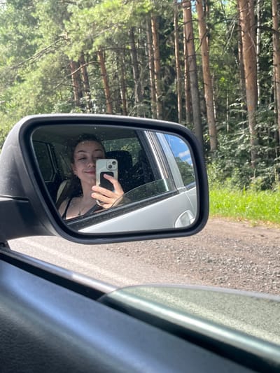 A young woman is reflected in a car's side mirror, taking a selfie with her phone. She is smiling and looking at the camera. She is wearing a black tank top and a necklace. The car appears to be on a dirt road in a wooded area, with tall pine trees visible in the background. The sky is blue with some clouds.