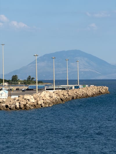 The image depicts a coastal scene at the Municipal Unit of Kastro - Kyllini, Greece. In the foreground, dark blue water with a rippled surface fills the lower half of the frame. A breakwater constructed of large, angular concrete blocks extends from the right side of the image towards the left.

Beyond the breakwater, a paved area appears to be a parking lot or loading zone. Several vehicles are parked, including cars and a large truck with a blue trailer. A white structure with a blue roof, resembling a small chapel, is visible on the far left. A tall pole with lighting fixtures is positioned on the left edge of the paved area.

Further into the midground, a line of four tall poles, each equipped with multiple lights, stretches across the scene. Behind these poles, lush green trees and vegetation line the coast. In the background, a large, hazy mountain range dominates the horizon, its slopes appearing a muted blue and gray against a clear, pale blue sky. A few wispy white clouds are scattered across the sky. The overall impression is a functional port area with a natural mountainous landscape in the distance.