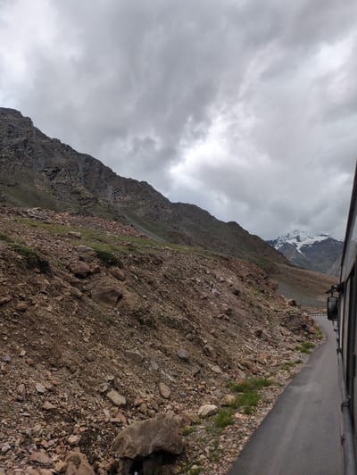 An overcast day captures a roadside view of a mountainous landscape near Zing Zing Bar, India. The foreground features a winding asphalt road on the right, with the side of a bus visible along its edge. The road curves away from the viewer, leading into the distance. The left side of the image is dominated by a steep, rocky mountainside, characterized by loose stones, gravel, and sparse patches of green vegetation. The mountain slopes upwards and recedes into the background. Further in the distance, snow-capped peaks are visible against a cloudy sky. The overall atmosphere is one of rugged, natural beauty under a somber sky.