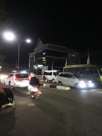 The image depicts a nighttime scene on a street in Walennae, Indonesia. A white SUV with its brake lights illuminated is in the foreground, with a person on a scooter behind it. To the right, a silver car is approaching with its headlights on. Further in the background, a yellow bus is visible, and behind it, a multi-story building with a distinctive triangular roof and illuminated windows. Streetlights are casting light on the road. The environment appears to be a moderately busy urban street at night.