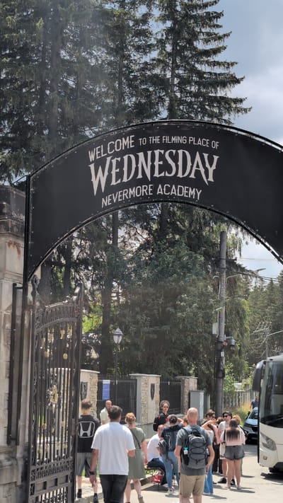 A group of tourists stands in front of a large, ornate gate at the entrance to a filming location in Bușteni, Romania. A black banner is suspended from an archway above the gate, which reads "WELCOME TO THE FILMING PLACE OF WEDNESDAY NEVERMORE ACADEMY" in white lettering. The gate is made of wrought iron and appears to be the entrance to a large estate or castle. The tourists are dressed in casual clothing and some are taking pictures. The background consists of tall evergreen trees and a blue sky. A white tour bus is parked to the right of the frame.