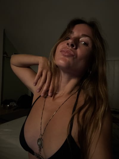 The image is a close-up portrait of a young adult woman, originally rotated 90 degrees clockwise. She looks directly at the viewer with a calm, slightly smirking expression. Her long, wavy light brown hair, streaked with lighter highlights, falls around her shoulders. A septum piercing is visible on her nose.
She wears a dark, thin-strapped top, possibly a bra or bikini top. A distinctive multi-strand metallic necklace is prominently displayed on her chest. It features a central round pendant with a pearl-like bead, flanked by intricate, feather-like designs. Her right hand is gently positioned on her left shoulder or collarbone area.
The background is dark and indistinct, suggesting an indoor setting. The lighting is dim, with an artificial light source illuminating her face from above and slightly to the side, creating soft highlights and shadows that accentuate her features. The overall mood is intimate and confident. No text is visible in the image. The location context for the image is Graz, Austria.