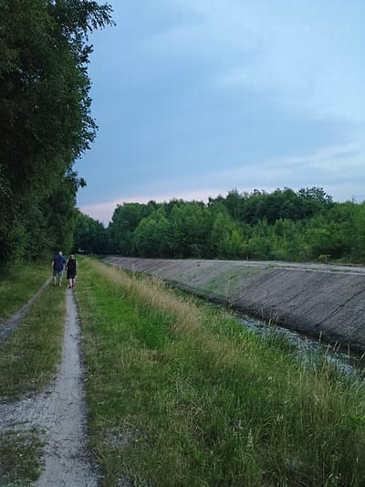 Two individuals are walking away from the camera on a dirt path beside a grassy area. The person in front is wearing a dark backpack and dark shorts with dark footwear. The person behind them is wearing a dark blue short-sleeved shirt and light gray shorts with dark footwear. To the right of the path, a steep, concrete-lined ditch with water at the bottom runs parallel to the path. Beyond the ditch is a dense line of green trees. To the left of the path, more trees with lush green foliage line the area, creating a natural border. The sky above is a pale blue with some clouds, indicating it is likely late afternoon or early evening. The overall setting appears to be a rural or park-like area. This location is identified as being in Papenburg, Germany.