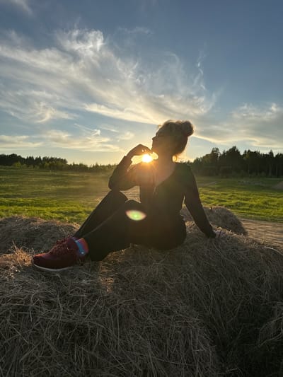 A silhouetted individual, likely a woman, is captured crouching or sitting on a large round hay bale in a rural field. The person is positioned in profile, facing left, with light-colored hair styled in a bun. She wears dark clothing, consisting of a long-sleeved top and pants, and burgundy sneakers with brightly colored striped socks visible on her ankles. Her right hand is raised towards the setting or rising sun, which is directly behind her head, creating a strong lens flare and backlighting effect that frames her silhouette and illuminates her hair.

The scene depicts a wide-open landscape under a vast sky. A large green field extends into the distance, bordered by a line of dark trees or a forest. The sky is a mix of deep blue and lighter hues, with elongated, wispy clouds. The low sun bathes the entire scene in a warm, golden glow. The presence of hay bales indicates an agricultural setting. The image evokes a serene and contemplative mood, set against the natural beauty of the countryside near Стеклянный, Russia. There is no visible text in the image.