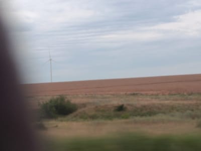 The image depicts a rural landscape in Sychavka, Ukraine, under a cloudy sky. In the background, a solitary wind turbine stands tall against the horizon, its blades blurred, suggesting it might be in motion or that the image is slightly out of focus. The middle ground is dominated by a large, reddish-brown field, likely cultivated land, with subtle horizontal striations that could indicate plowing or natural contours. In the foreground and lower middle ground, there is an expanse of dry, grassy terrain interspersed with patches of green vegetation, including a small cluster of trees or bushes. The overall scene conveys a sense of open space and agricultural activity.