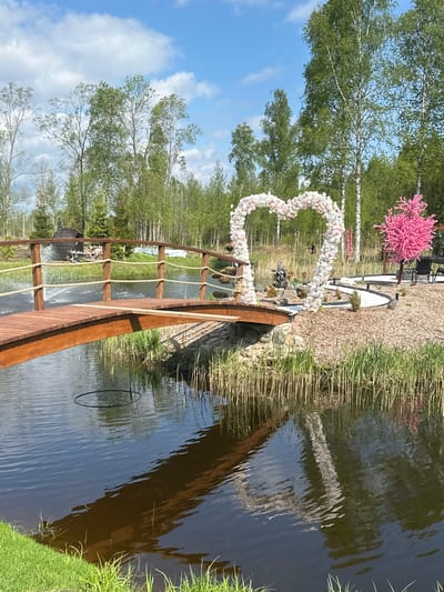 The image shows a picturesque outdoor scene, likely a wedding venue, in Vaibla, Estonia. A heart-shaped floral archway stands near the edge of a pond or lake, adjacent to a wooden bridge with rope railings. The water reflects the bridge and surrounding vegetation. The backdrop includes trees, including what appears to be a pink cherry blossom tree. In the distance, some outdoor seating and other decorative elements are visible. The sky is blue with scattered clouds.