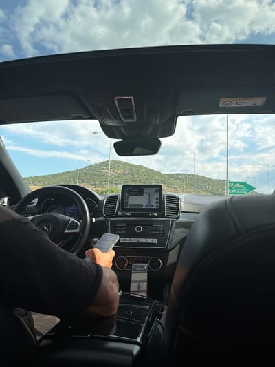 The image, taken from the passenger seat of a car, shows the interior dashboard and the view through the windshield. The driver's hands are visible on the steering wheel and holding a smartphone. The car's navigation system is displayed on a screen, showing a route with "129 km" remaining. In the background, a green road sign indicates "Exit Kavala". The landscape outside features a green, hilly terrain under a cloudy blue sky, with streetlights visible along the road. The overall scene suggests a road trip or commute within or near Agios Andreas, Greece, given the presence of Greek text on the road sign.