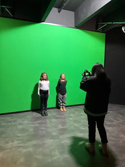 This image depicts an indoor photography session in a studio located in Canela, Brazil. Two children, appearing pre-adolescent, stand side-by-side facing forward in front of a large, illuminated green screen that extends from floor to ceiling. The child on the left wears a white long-sleeved shirt, black pants, and dark boots. The child on the right wears a dark long-sleeved shirt, grey patterned cargo pants, and light brown boots. Both children are smiling faintly. In the foreground, an adult, viewed from the back, holds a black DSLR camera aimed at the children. This adult wears a dark jacket with a white band at the lower hem, dark pants, and light-colored boots. The studio environment features dark, possibly black, walls and ceiling, with visible dark structural elements and pipes overhead. The floor is composed of large, mottled grey square tiles. Shadows from the subjects are cast onto the tiled floor directly in front of the green screen.