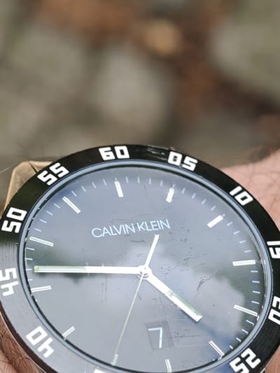 A close-up, high-angle, forensic view in Tsarev Brod, Bulgaria, captures a dark-faced "CALVIN KLEIN" wristwatch secured to an arm, with visible arm hair around the casing. The watch face is significantly scratched, particularly across the center. The minute hand points directly to the 45-minute marker, the hour hand rests between the 8 and 9 o'clock positions (closer to 9), and the second hand indicates the 30-second mark, showing an approximate time of 8:47:30. A date window at the 6 o'clock position displays the number "7". The black bezel features white numerical markers at five-minute intervals from "05" to "60". The background is an indistinct, out-of-focus blur of natural earth tones and greens, suggesting an outdoor environment.