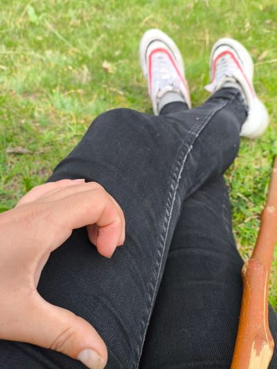 A first-person perspective shot shows a person sitting outdoors on a grassy area. The person is wearing black jeans and white sneakers with red accents. Their left leg is crossed over their right, and their left hand is resting on their left thigh, forming a heart shape. To the right of the frame, a brown wooden stick is visible, resting against the person's legs. The background consists of out-of-focus green grass.