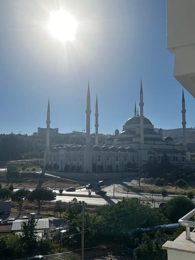 The image captures a wide view of the Kocatepe Mosque in Ankara, Turkey, bathed in bright sunlight under a clear blue sky. The grand mosque, with its numerous minarets and a large central dome, dominates the center of the frame. It is situated on a hill, with a road and some vehicles visible in the foreground. The architecture of the mosque is characterized by its intricate detailing and white marble facade. The surrounding landscape includes trees and hills, with a few modern buildings visible in the background. The sun is positioned in the upper left corner of the image, creating a lens flare effect. The overall scene depicts a serene and majestic view of the mosque on a sunny day.