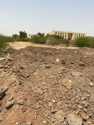 The image shows a pile of dirt and rubble in front of a large building in Khartoum, Sudan, located at the coordinates 15.528 latitude, 32.571 longitude. The dirt is a mix of brown, gray, and red, with some small rocks visible. The building is a large, rectangular structure with a flat roof. There is a small amount of vegetation in front of the building, and the ground is dusty and dry.  The building appears to be unfinished, as there are no windows or doors. The foreground is dominated by the dirt pile, with the building in the background. There are no people visible in the image.