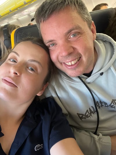 Two individuals, a male and a female, are depicted in a close-up selfie taken inside a commercial aircraft cabin, likely at or near Girona-Costa Brava Airport (GRO), serving Vilobí d'Onyar, Spain. The male, positioned on the right, is a Caucasian individual appearing in his 30s-40s, with short grey hair, blue eyes, light complexion, and stubble. He is smiling broadly and wears a light grey hooded sweatshirt with a visible black drawstring and a dark, partially obscured graphic design on the chest. The female, positioned on the left and slightly closer to the camera, is a Caucasian individual appearing in her 20s-30s, with long dark brown hair, blue eyes, and a light complexion. She has a small stud earring in her right ear. She wears a dark blue collared polo shirt featuring a white Lacoste crocodile logo on the left chest. Her head is tilted, resting against the male's left shoulder/upper arm, and she looks towards the camera with a neutral expression. The background reveals typical airplane seating with yellow accents on the headrests and light-colored overhead panels, with other blurry passengers visible.