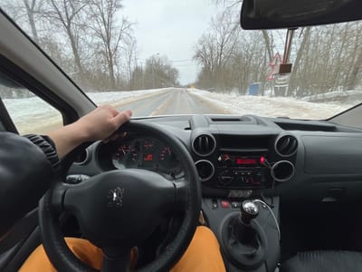 The image captures a driver's first-person perspective from inside a Peugeot vehicle on a winter road in Rogachovo, Russia.
**Interior:** The driver's right arm and hand, clad in a dark sleeve, firmly grip the top of the black, possibly covered, steering wheel featuring the Peugeot lion emblem. Below the steering wheel, the driver's lap is visible, covered by orange fabric trousers. The dark grey dashboard is equipped with circular air vents, a central infotainment system displaying red text, and various control buttons, including a hazard light switch. A red-tipped cable extends from the dashboard's lower right. To the driver's right, a manual gear shifter with a metallic knob is prominent. The instrument cluster behind the steering wheel shows partially visible gauges with red needles and a digital display. A rearview mirror is affixed to the windshield, from which a small, reflective, potentially triangular object hangs.
**Exterior (seen through windshield):** The vehicle is on a two-lane road that appears wet or slushy, with a faint yellow centerline. Significant banks of dirty snow and ice line both sides of the road. Beyond the snow, bare deciduous trees form a dense line, characteristic of winter. The sky is overcast and uniformly grey. On the right side of the road, further down, a small blue portable structure is visible behind the snowbank, accompanied by a red-bordered triangular warning sign (potentially indicating uneven road or other hazards) and utility poles with power lines. The overall environment suggests cold, wintery conditions.