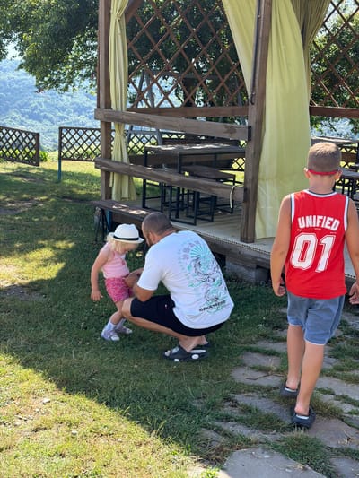 In Izmaylovka, Russia, an outdoor scene unfolds with a man crouching and interacting with a young girl who is wearing a white hat and a pink top. The man is dressed in a white t-shirt and black shorts. Behind them, a boy in a red basketball jersey and blue shorts walks away from the camera, facing a wooden gazebo. The gazebo has a lattice design and is partially covered by light yellow curtains. In the background, lush green trees and a hilly landscape suggest a rural or semi-rural setting. The ground is covered with grass and a stone pathway. The overall impression is a family enjoying a sunny day outdoors.