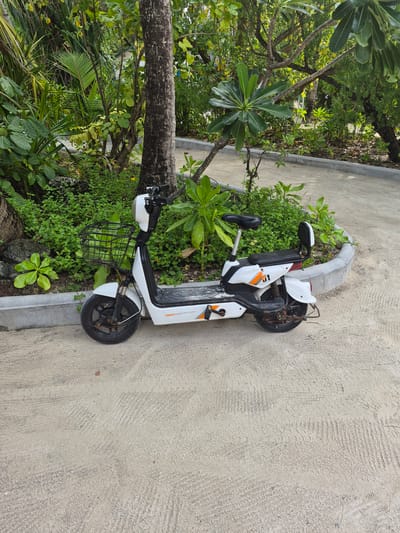 The image shows a white electric scooter parked on a sandy pathway in Huraa, Maldives. The scooter has a black basket in the front and an orange accent on the body. It is leaning against a concrete curb that separates the path from a patch of green vegetation, including trees and bushes. The pathway appears to be sandy, with tire tracks visible. The scene suggests a tropical, outdoor environment.
