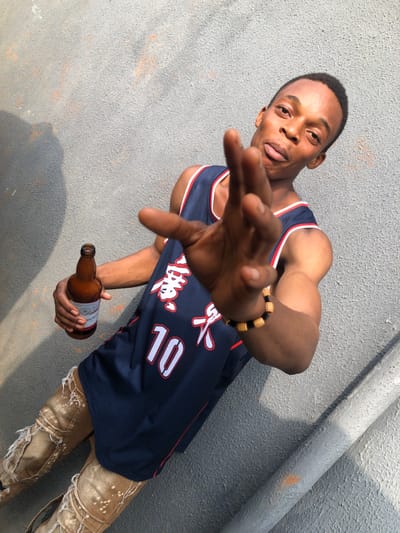 A young man in a dark blue basketball jersey with the number 10 poses with a hand outstretched. He wears a beaded bracelet and ripped, light-colored jeans. He holds an amber beer bottle in his other hand. The background shows a gray wall and part of a pipe. The image has a strong light source casting shadows on the wall and the man.