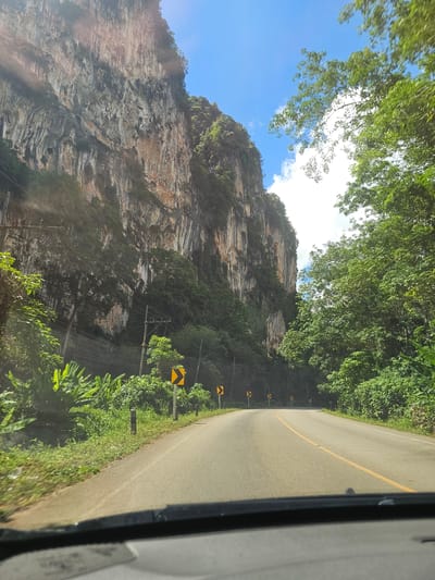 The media file shows a view from a vehicle traveling along a road in Ban Nai Sa, Thailand. The scene is dominated by a towering, sheer rock face on the left, characterized by its striated texture and patches of greenery clinging to its surface. Lush green foliage, including trees and banana plants, lines the right side of the road, creating a tunnel-like effect with branches extending overhead. The road itself is paved with a yellow line marking the center. Yellow warning signs with black arrows, indicating curves in the road, are visible along the roadside. The sky above is a bright blue, with scattered white clouds. The sunlight suggests it is daytime, likely morning or afternoon, and the weather appears clear. The perspective implies the vehicle is in motion, possibly on a scenic drive through a natural landscape. There is no visible text within the image.