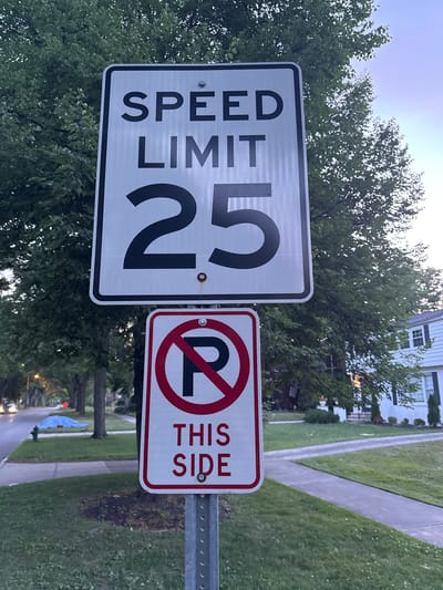 This is a photograph of a street sign in Shaker Heights, Ohio, United States. The sign indicates a speed limit of 25 mph. Below the speed limit sign, there is another sign with a red circle and slash over a black "P" symbol, indicating "no parking." This sign also reads "THIS SIDE." The signs are mounted on a metal pole. The background consists of trees, a sidewalk, a street, and a residential house. The sky is visible in the upper right portion of the image.